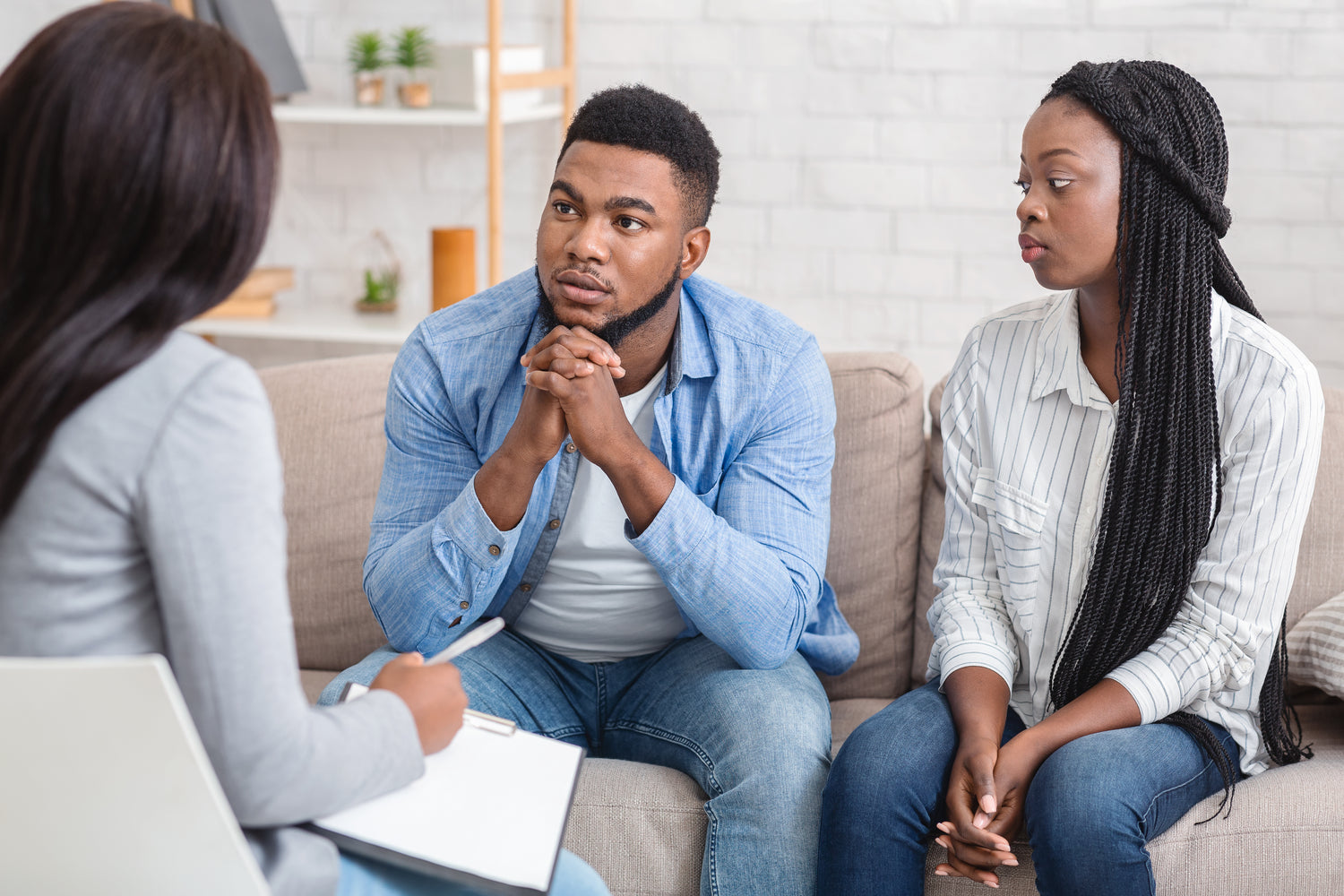 A couple sitting on a couch during a therapy session, attentively listening to their therapist. The man sits with his hands clasped, while the woman looks thoughtful. The therapist takes notes, facilitating a supportive conversation in a comfortable setting, focusing on mental health care and relationship counseling.