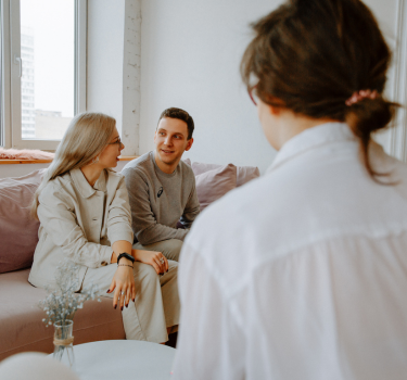 Two people sitting on a couch during a couples therapy session, attentively listening to their therapist, who is holding a tablet. The session takes place in a modern, calm setting with natural lighting, focusing on relationship counseling and emotional support.