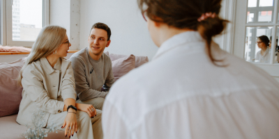 Two people sitting on a couch during a couples therapy session, attentively listening to their therapist, who is holding a tablet. The session takes place in a modern, calm setting with natural lighting, focusing on relationship counseling and emotional support.