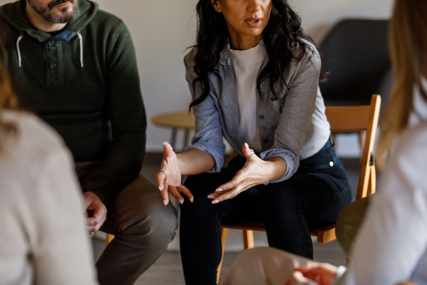 Group therapy session where a female participant speaks with her hands gesturing while others listen attentively. The group is seated in a circle, fostering a supportive and open environment for mental health discussions.