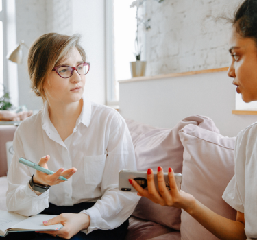 Therapist and client engaging in a one-on-one counseling session. The therapist takes notes while the client speaks, appearing open and expressive. They are seated in a bright room with large windows, creating a comfortable and welcoming environment for mental health care and therapy.