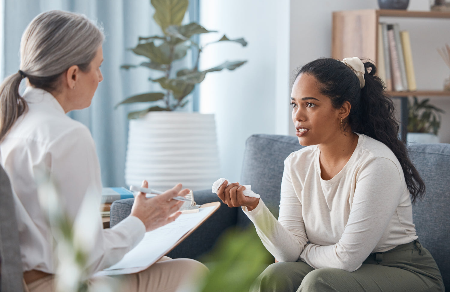 Female therapist providing counseling to a young woman in a therapy session. The woman, holding a tissue, expresses her feelings while the therapist listens attentively and takes notes. The scene takes place in a calm, supportive environment, emphasizing mental health care, therapy services, and emotional support.